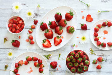Top view of fresh strawberry whole and cutted in bowls and on plates decorated with flowers over white table.