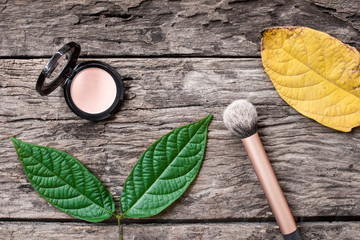 women cosmetic cheek powder and brush with green and yellow leaves on wooden background