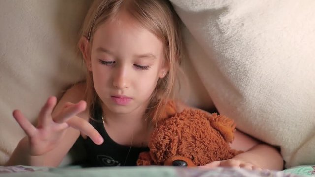 Cute little girl sitting on bed together with teddy bear playing games on tablet.