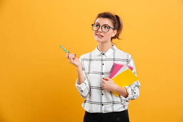 Photo of young woman looking aside while standing with textbooks and pen in hands, isolated over...