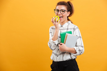 Photo of happy smiling woman posing at camera with books in hands and eating ripe green apple, isolated over yellow background