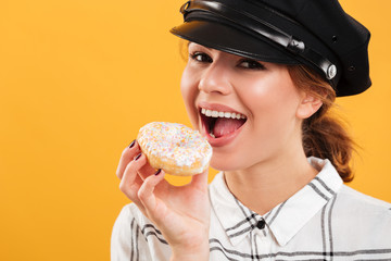 Portrait closeup of joyful woman in plaid shirt and police hat eating sweet delicious donut, isolated over yellow background