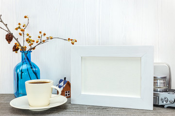 empty white photo frame, cup of coffee, blue glass vase with dried flowers on wooden background