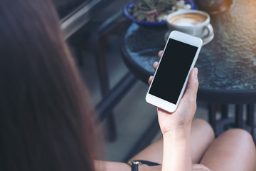 Mockup image of a woman's hand holding white mobile phone with blank black desktop screen and coffee cup in cafe