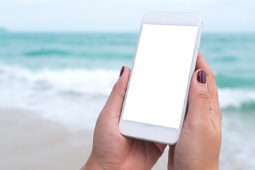 Mockup image of woman's hands holding white mobile phone with blank desktop screen in front of the sea and blue sky background