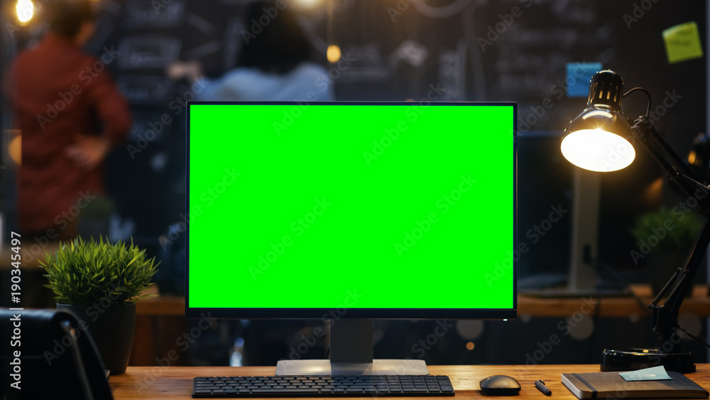 Poster personal computer with mock-up green screen monitor stands on the office desk, in the background peo