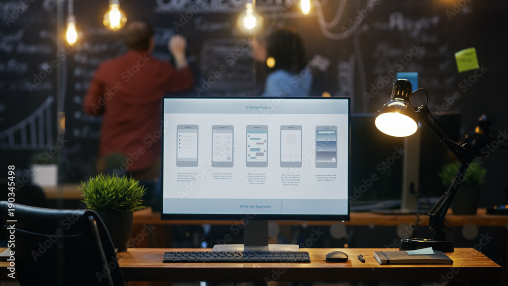 Poster Personal Computer with Mobile Application Design Showing on the Monitor Stands on the Office Desk, In the Background People Work on a Blackboard Wall in the Creative Office.