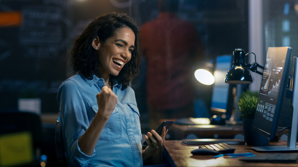 Beautiful Female Financier Uses Mobile Phone, Has Great News and Shows "Yes" Gesture, Her Personal Computer Showing Graphs and Statistics. She Smiles Charmingly.