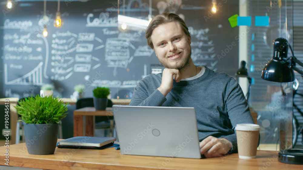 Poster Handsome Young Office Employee Thinks on a Problem Solution While Typing on a Laptop Computer,  He Charmingly Smiles on Camera. He's Working in the Creative Stylish Office.