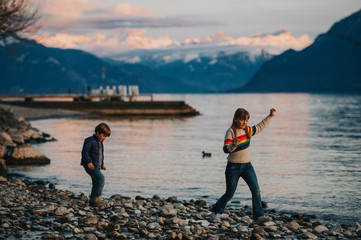 Two happy kids, little brother and big sister, playing together by lake Geneva at sunset with swiss mountains Alps on background. Image taken in Lausanne, Switzerland