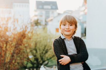 Outdoor portrait of cute 5 year old blond boy, wearing grey polo shirt and black knitted jacket