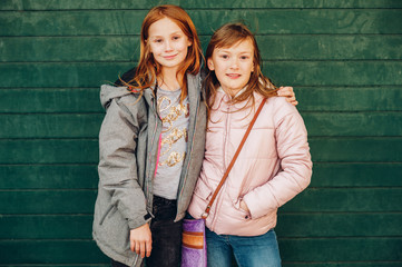 Outdoor portrait of two cute little teen girls wearing warm jackets, posing against green background, fashion for children