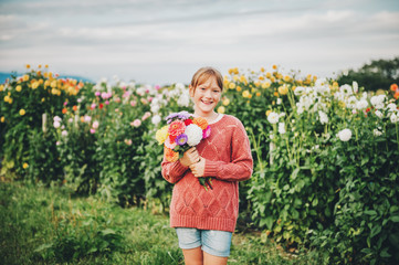 Outdoor portrait of cute little girl holding colorful bouquet of dahlia flowers, wearing fashion pullover