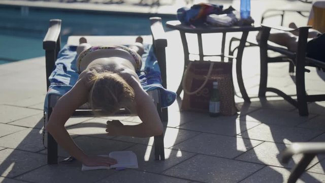 Medium shot of woman with her straps undone for her bikini top, lying on a chaise lounge and reads her book resting on the ground.