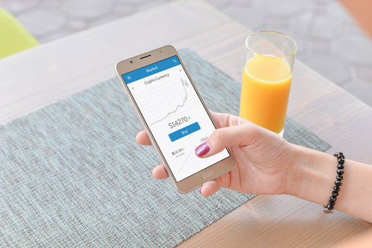Crypto Currency Graph On Smart Phone App. Woman Holding Phone. Juice And Table In Background.