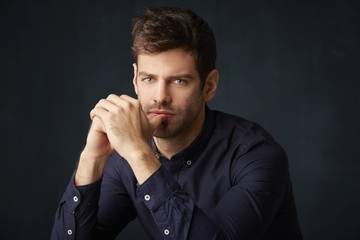 Thinking young man studio portrait. Studio portrait of a thinking young man with hands at his face looking at camera while sitting at dark background. 