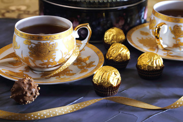 Tea ceremony, tea party. Two tea cups of gold color with black tea, candy, chocolate and a box with cookies on a lilac-colored background.