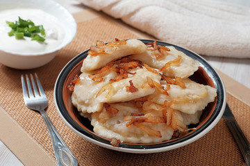 A plate with boiled dough vareniki with potato filling and fried onions. Near her bowl with sour cream, fork and knife.