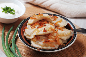 A plate with boiled dough vareniki with potato filling and fried onions. Near her bowl with sour cream, fork and greens.