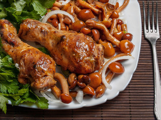 Chicken drumstick baked in the oven lies on a white plate with pickled mushrooms and greens