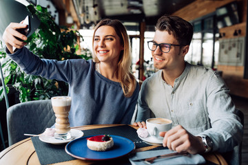 Pretty girl taking a selfie with her boyfriend, using smartphone, while spending time at cafe with cup of coffee.