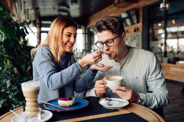 Beautiful smiling girl feeds her handsome boyfriend, eating tasty cake and drinking coffee, spending time together at cafe.
