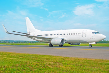 White passenger airplane rides the track taxiway at the airport, against a background of a blue sky in the foreground a green lawn.
