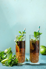 Two Glasses of classic Cuba libre cocktail with lime, mint, crushed ice and cola, served with ingredients above and retro cocktail tubes over turquoise green pin-up style background. Sunlight, space