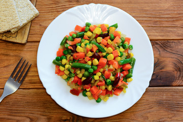 Mixed vegetable stew on a serving plate and wooden table. Homemade stewed carrots, green beans, red beans, peas, corn, pepper and onions. Healthy vegetarian stew. Top view