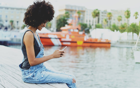 Model looking dark-skinned girl with afro hair typing text message on a modern cellphone while sitting on a wooden embankment on a yacht background. Smiley female using smartphone for check e-mail.