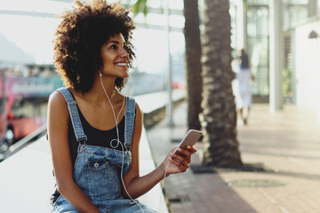 a happy dark-skinned girl enjoys music from her playlist through headphones. girl uses a mobile phone to watch a new video of her favorite artist. Female person listening online radio via cellphone