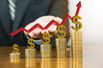 Businessman hand holding red arrow up with golden dollar sign on money coin stack arranged as a graph on wooden table, concept of money and business growth