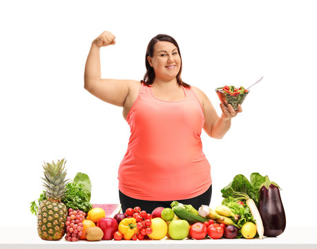Overweight woman holding a bowl of salad and flexing her biceps behind a table with vegetables and fruit