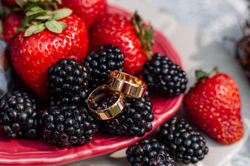 Wedding gold rings, laying ceramic dishes with plums, strawberries, blackberries. Bridal jewellery details and decorations with fruits