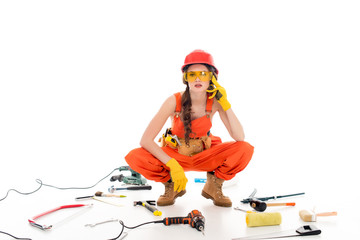 workwoman in overalls sitting on floor with different equipment and tools, isolated on white