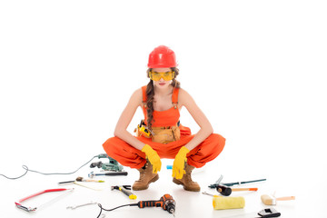 confident workwoman in overalls sitting on floor with different equipment and tools, isolated on white