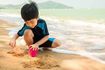 A boy in swimming suit is sitting on the beach and playing sand at the sea in the sunny day in summer