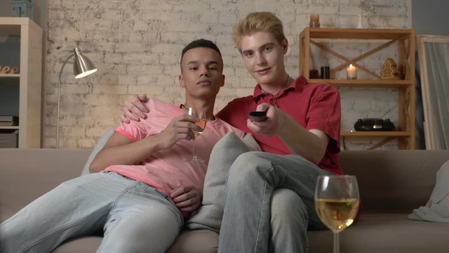 Multinational gay couple sitting on couch, watch funny TV show, laughing, use the remote control, look at the camera. Homeliness, romantic evening, hugs, happy LGBT family concept. 60 fps