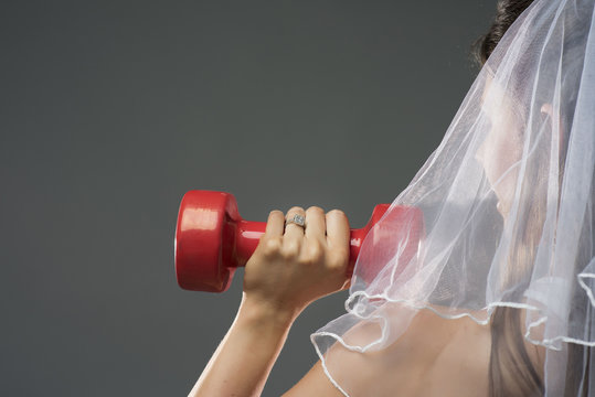 closeup rear view of a  young woman head wearing a white wedding bridal veil holding red dumbbells on a grey background in studio 