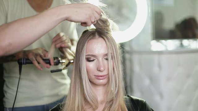 A man winds the hair of a young blonde model with a curling iron in a beauty salon.