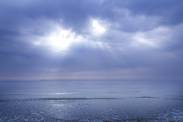 Obraz premium Sun shining through thick clouds of overcast cloudy sky caused ray of light beaming down to the blue ocean surface
