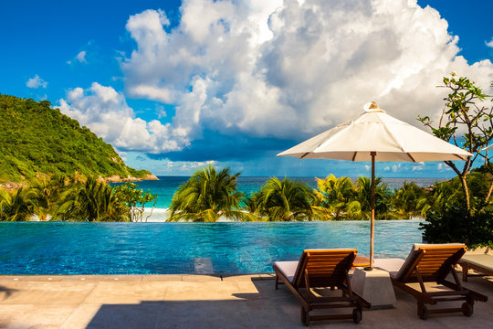 A great holiday image of two deck chairs next to each other, shaded by a patio umbrella in between and in front is a wonderful view of an infinity pool and the beach.