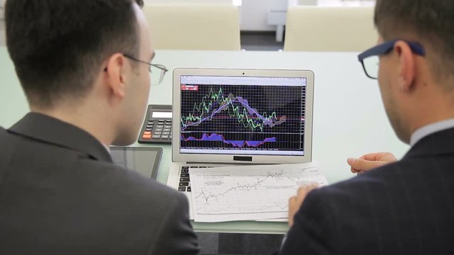 The two finance analytics work on the final report using the brand new laptop and draft. The colleagues look at the charts on the screen and compare the data with information on the paper. Men wears