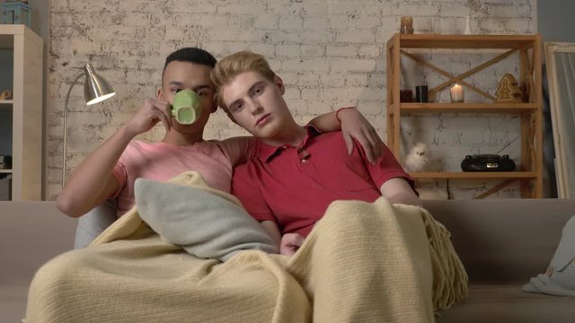 Multinational gay couple sitting on couch covered with a warm blanket, watch TV, use the remote control, look at the camera. Homeliness, romantic evening, cuddles, happy LGBT family concept. 60 fps