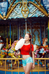 Fototapeta na wymiar Young beautiful sexy woman in a red virgin t-shirt glasses blue shorts white sneakers eating sweet cotton wool in the summer in the park carousel