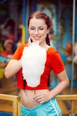 Young beautiful sexy woman in a red virgin t-shirt glasses blue shorts white sneakers eating sweet cotton wool in the summer in the park carousel