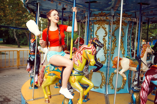 Young beautiful sexy woman in a red vest with pigtail glasses blue shorts white sneakers eating sweet cotton wool in the summer in the park and riding a carousel horse