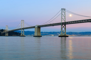 San Francisco-Oakland Bay Bridge with full moon rising and pink and blue sunset skies. The Embarcadero, San Francisco, California, USA.