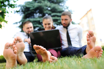  friends in business clothes and barefoot sitting on grass holding laptop in hands