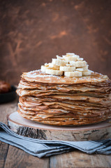 On the table on a stand made of wood cut a pile of thin pancakes, sprinkled with jam, sprinkled with powdered sugar, on top are banana and peanuts. In the background a plate with dates. Style Rustic